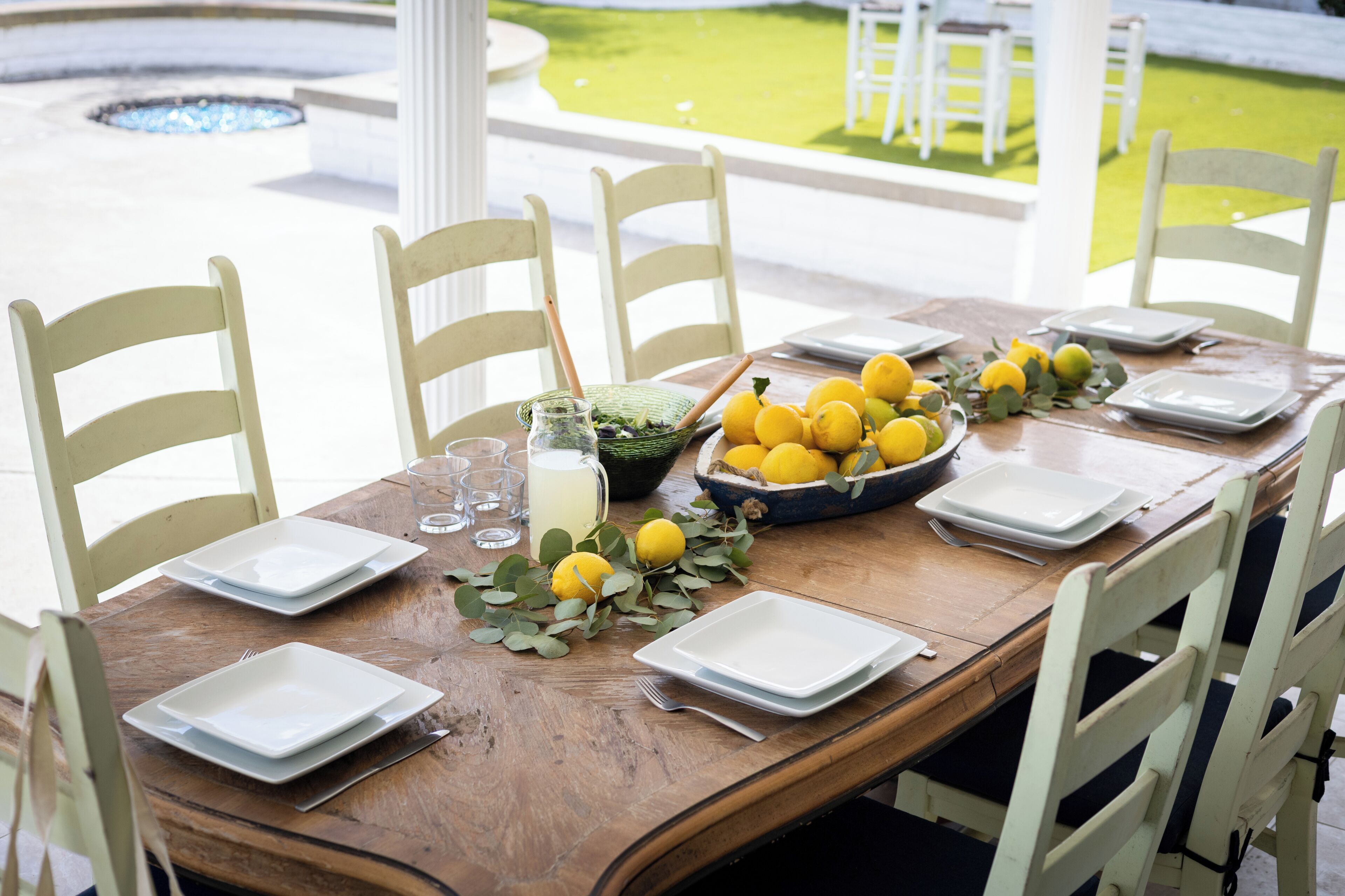 Mesa de comedor dispuesta para seis personas, con platos blancos, un cuenco de limones y una jarrón de limonada.