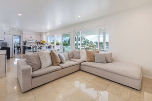Ein modernes Wohnzimmer mit einem beigefarbenen Ecksofa, einem Essbereich mit weißem Tisch und einer Küche im Hintergrund.