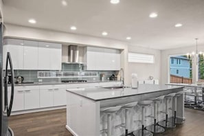 A modern kitchen with white cabinetry, a central island with bar stools, a stainless steel range hood, and a large window.