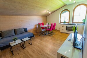 Een knus ingerichte kamer met houten plafond, een bank, een eettafel met stoelen, en een keukenhoek met een fornuis en ramen.
