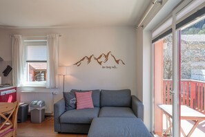Ein Wohnzimmer mit grauem Sofa, einem Fenster mit Vorhängen, einem Heizkörper, einem Mülleimer und Blick auf einen Balkon mit roter Brüstung.