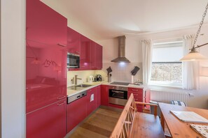 Eine kleine Küche mit roten Schränken, einem Holzespeisetisch, einem eingebauten Backofen und einem Fenster mit Vorhängen.
