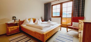 Ein Schlafzimmer mit einem hölzernen Bett, einem Nachttisch, einem Stuhl, einer Holzkommode und einem großen Fenster mit Vorhängen.