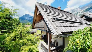 Eine traditionelle Holzhütte mit geneigtem Dach, umgeben von üppiger Vegetation und Bergen im Hintergrund.