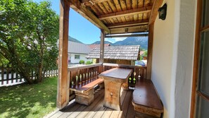 Eine Holzterrasse mit Bank und Tisch, eine weiße Wand und Blick auf Häuser und Berge.