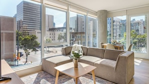 A modern living room with a sofa, a coffee table, and a view of the cityscape through large windows. - Rooftop Pool + Spa + Sky Lounge | Little Italy + Bay Views | Ancora by AvantStay (San Diego)