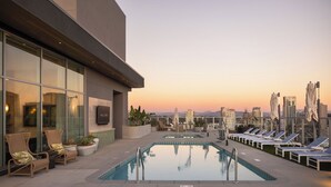 Uma piscina no terraço com espreguiçadeiras, uma parede de vidro e vista para a cidade ao pôr do sol.