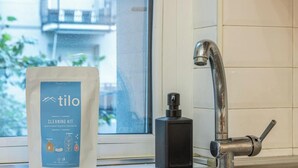 Ett fönster med utsikt över byggnader, en modern blandare och en väska med städmaterial.