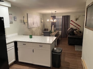 A kitchen with white cabinets, a granite countertop, a dining area with a table and chairs, a black leather sofa, and a chandelier.