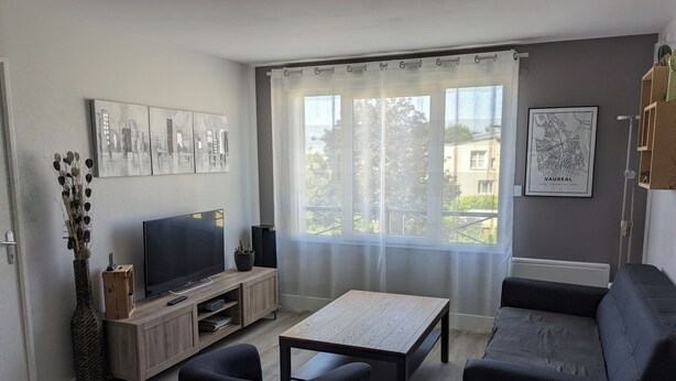 Ein modernes Wohnzimmer mit Flachbildfernseher, einer Holz-Unterhaltungsecke, einer Couch, einem Couchtisch und einem großen Fenster mit transparenten Vorhängen.