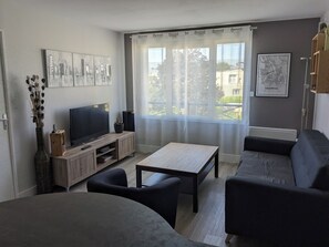 Ein modernes Wohnzimmer mit Flachbildfernseher, einer Holz-Unterhaltungsecke, einer Couch, einem Couchtisch und einem großen Fenster mit transparenten Vorhängen.