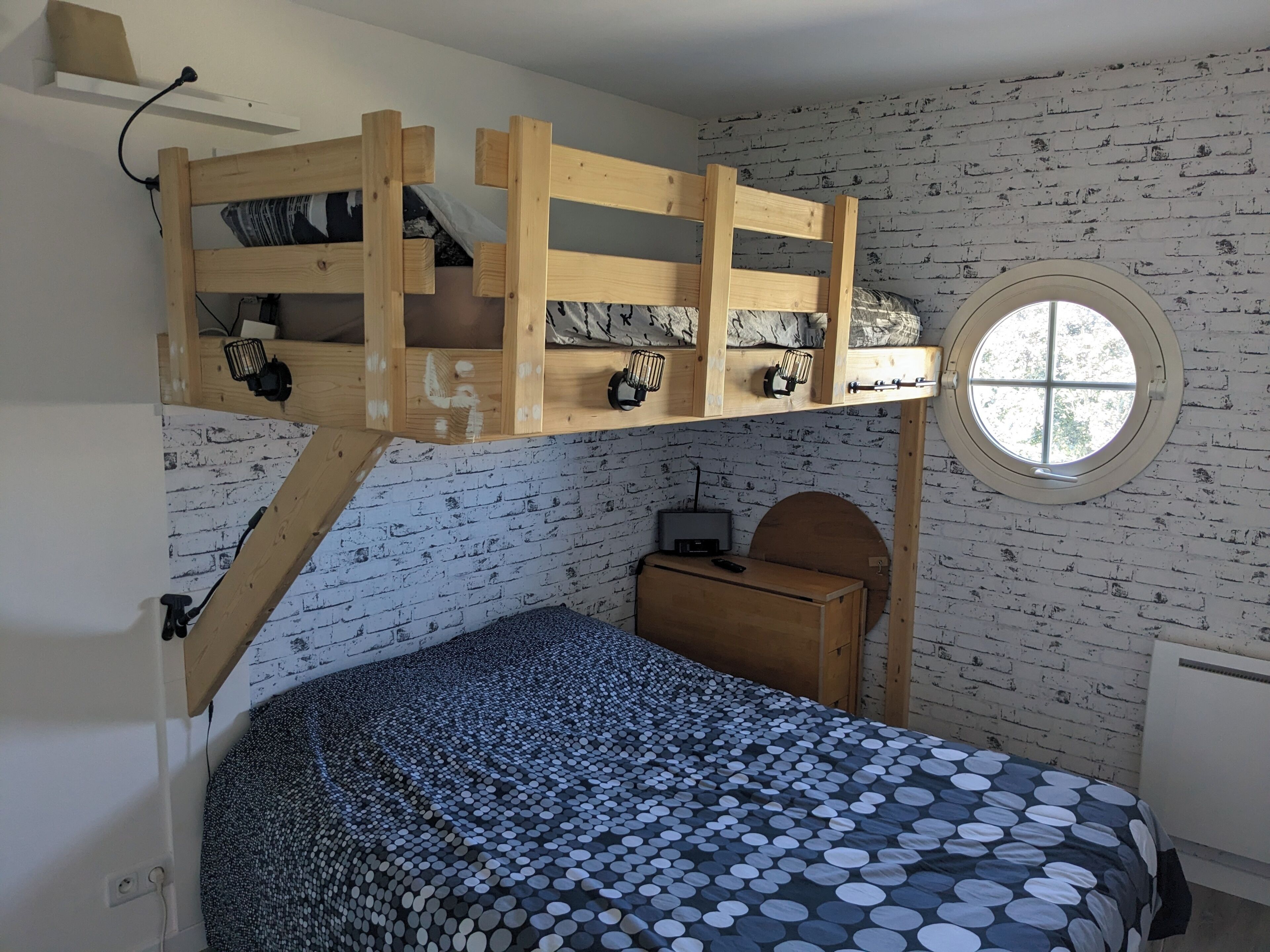 A bunk bed with a desk and a round window in a room with a brick-patterned wall.