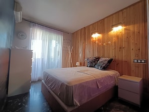 A bedroom with a wooden wall, a bed, a bedside table, and a window with curtains.