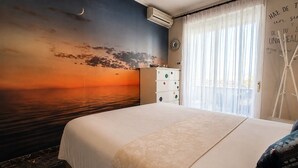 Ett hotellrum med en stor säng, en byrå, en väggmonterad luftkonditioneringsanläggning och utsikt över havet vid solnedgången.