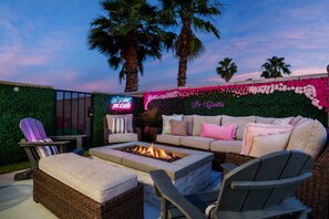 Area esterna con posti a sedere, un braciere, mobili in vimini e un muro decorato con fiori e insegne al neon.