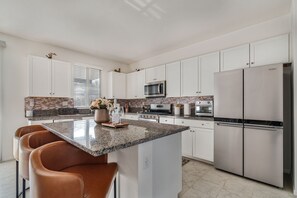 Una cocina moderna con gabinetes blancos, encimeras de granito, una isla central con asientos, electrodomésticos de acero inoxidable y un salpicadero de ladrillo.