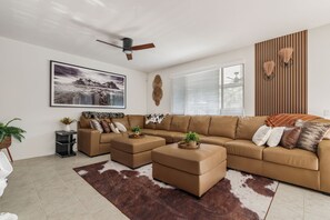 A modern living room with a sectional sofa, ottomans, a ceiling fan, a large framed picture, and a window with blinds. - Pool + Putting Green + Game Room | Coachella Fest | La Casita Retreat Coachella (Coachella)