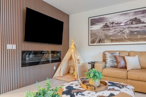 A modern living room with a flat-screen TV mounted on a wooden wall, a cozy sofa with cushions, a decorative teepee, and a mounted artwork of a mountainous landscape.