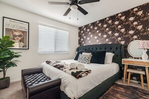 A bedroom with a bed, a ceiling fan, a potted plant, a bench, a desk with a mirror, and a floral wallpaper. - Pool + Putting Green + Game Room | Coachella Fest | La Casita Retreat Coachella (Coachella)