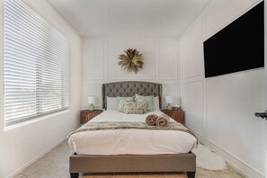 Camera da letto con testiera trapuntata, un grande elemento decorativo a parete, un letto con asciugamani arrotolati e una finestra con tende.