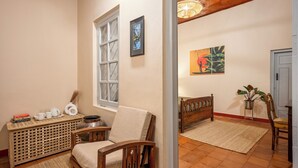 Quarto com uma cadeira de madeira, uma mesa pequena com livros e um vaso, uma janela com vista e um quadro na parede.