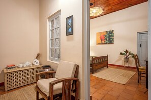 A room with a wooden chair, a small table with books and a vase, a window with a view, and a painting on the wall.