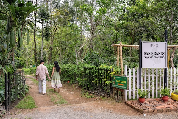 A pathway leading to a sign for 'RAHO SAND BANKS' with a white picket fence and potted plants.