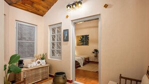 Quarto aconchegante com teto de madeira, uma janela grande, uma mesa de cabeceira com uma planta e uma cama com cobertor branco.