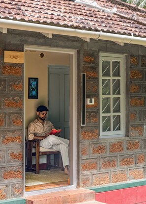 A man sitting on a chair outside a building with a tiled roof and a window with a grid pattern.