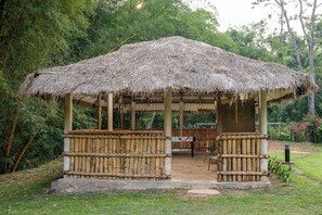A thatched-roof pavilion with a wooden bench and a table, surrounded by greenery.