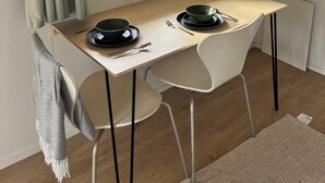 Spiseområde med et bord dækket til to, et vindue med gardiner og udsigt til grøn vegetation udenfor.