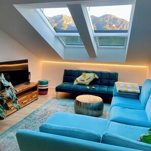 Ein Wohnzimmer mit einem blauen Sofa, einem Fernseher und einem Oberlicht mit Blick auf Berge.