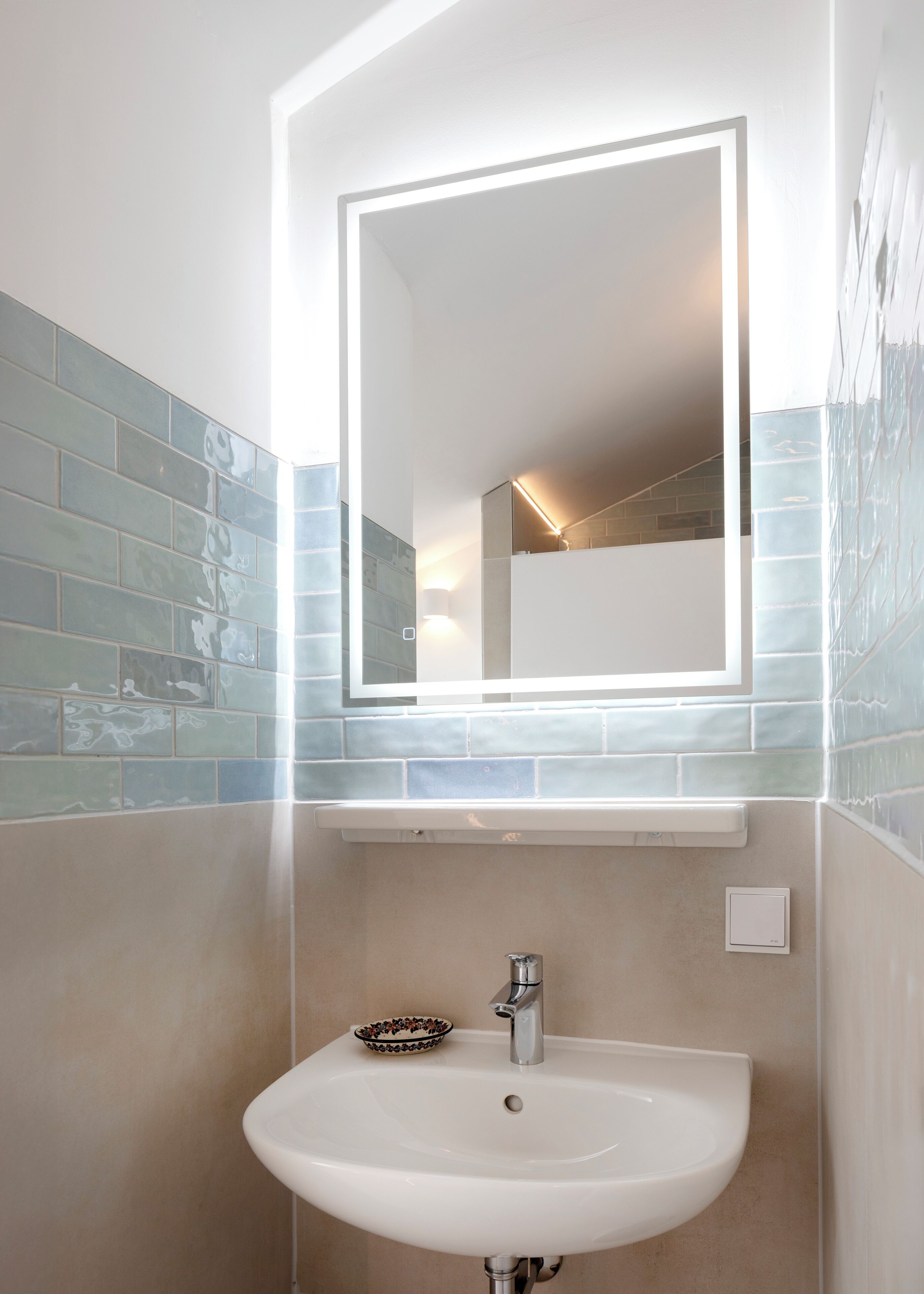 A bathroom with a white sink, a mirror with integrated lighting, and blue-tiled walls.