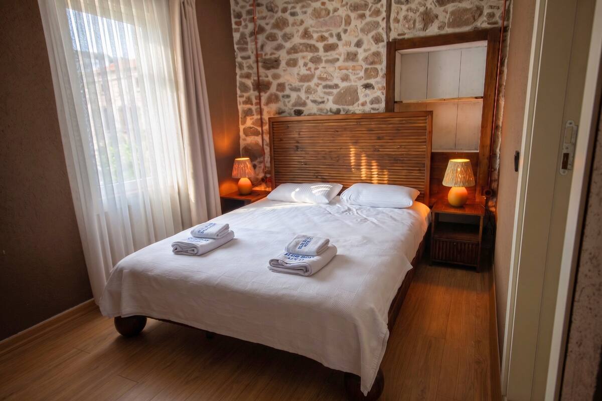Habitación de hotel con pared de piedra, cabecera de madera, cama con ropa blanca, dos lámparas de noche y piso de madera.