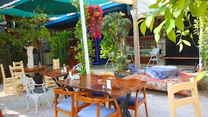 Un área de comedor al aire libre con mesas y sillas de madera, rodeada de follaje y cubierta por un toldo azul.