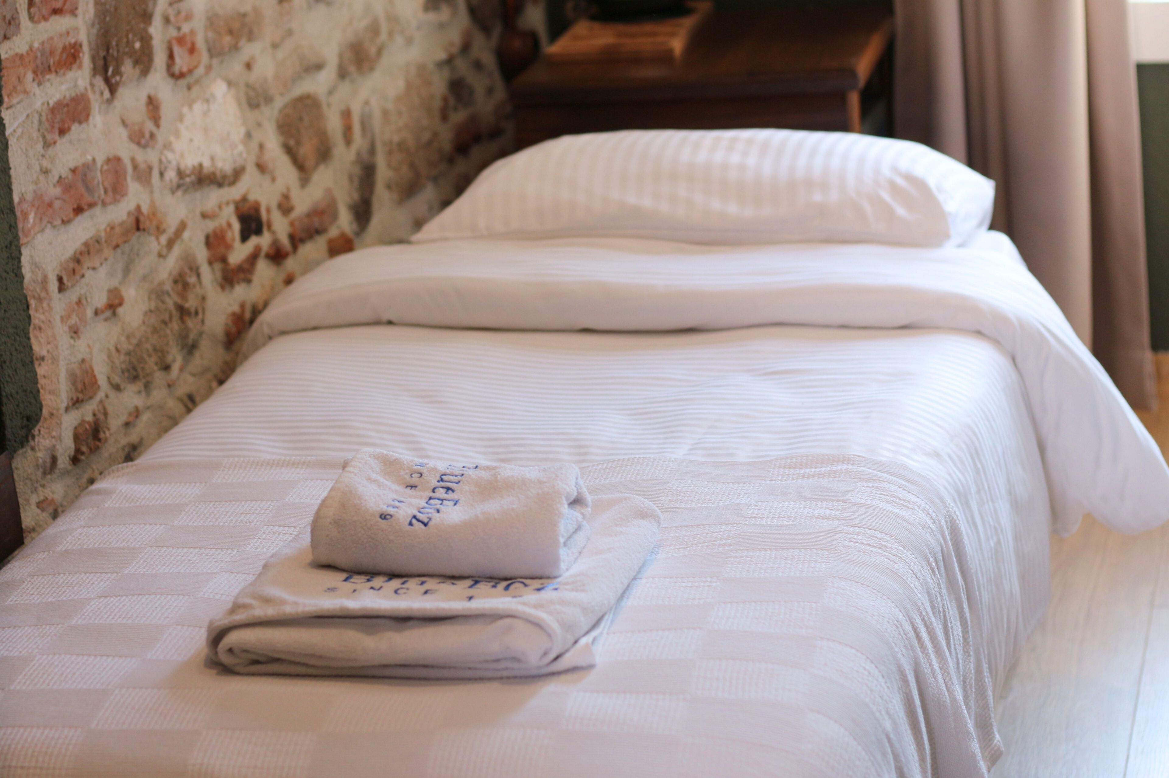 Un letto rifatto con un asciugamano piegato e un cuscino, una parete in mattoni e un comodino in legno.