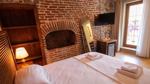 Habitación de hotel con chimenea de ladrillo, una cama con ropa blanca, una mesita de noche con lámpara y una ventana con cortinas.