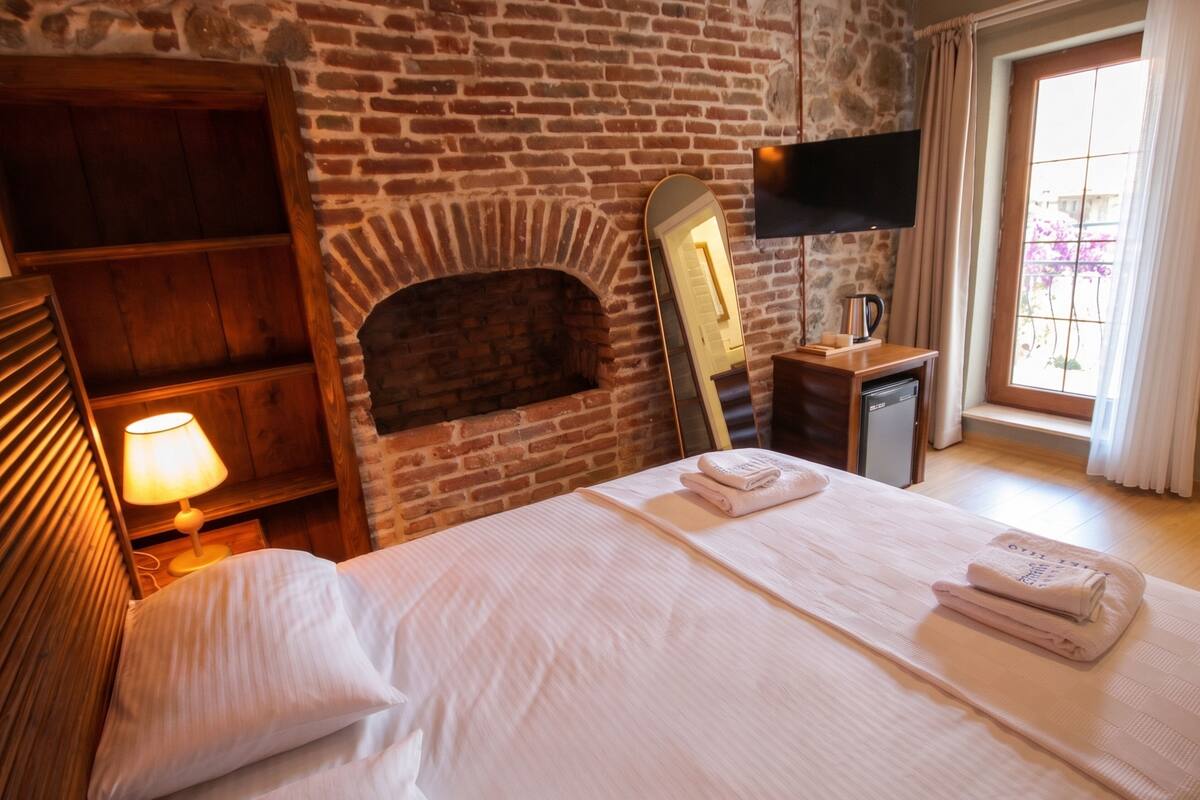 Habitación de hotel con chimenea de ladrillo, una cama con ropa blanca, una mesita de noche con lámpara y una ventana con cortinas.