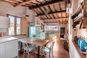 Eine rustikale Küche mit Holzbalken, einem Esstisch mit Stühlen, einem Kühlschrank und Blick durch ein Fenster.