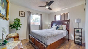 Un dormitorio con cama de madera, ventilador de techo, ventana con cortinas, mesita de noche con lámpara y una planta.