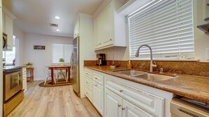 Una cocina con armarios blancos, encimera de granito, pileta de acero inoxidable y un comedor con mesa y sillas.