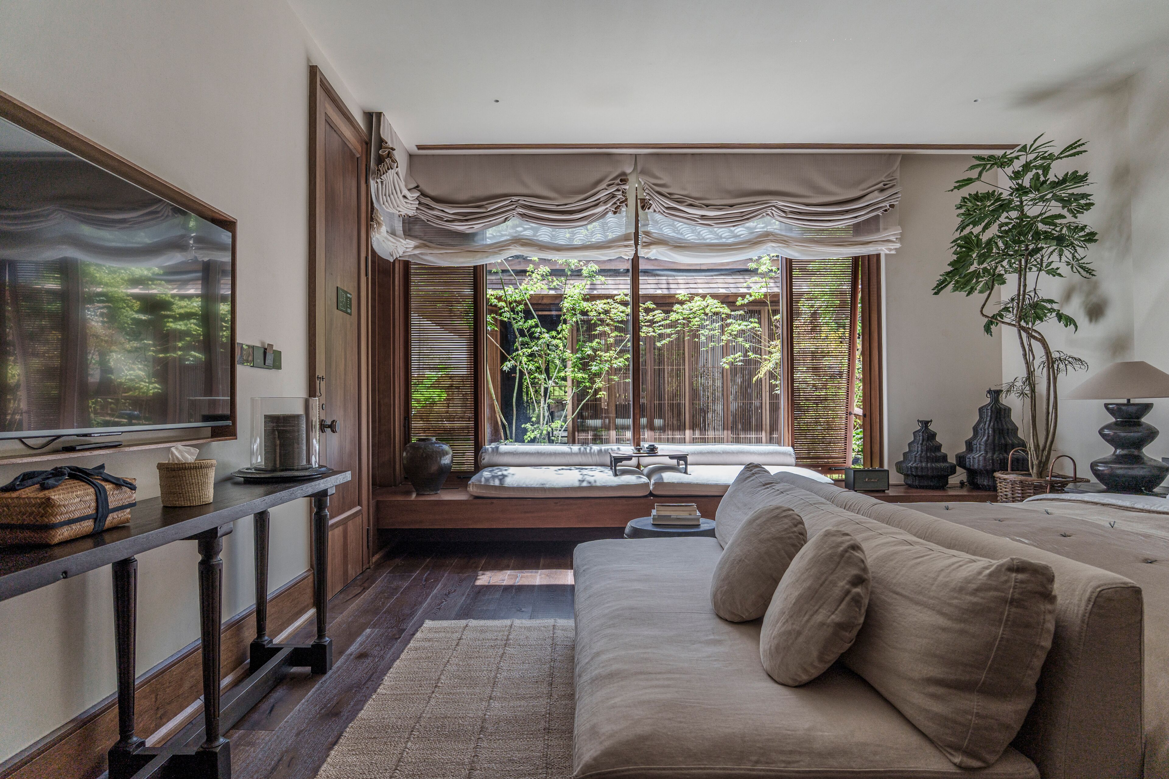A modern living room with a sofa, a television, and a view of a garden through large windows.