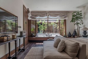 A modern living room with a sofa, a television, and a view of a garden through large windows. - MistyHues DawnGreen (Lijiang)