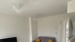 A modern living room with a flat-screen TV, grey sofas with yellow cushions, a chandelier, and a white door leading to another room. - House Sleeps 5 With Balcony (Hull)