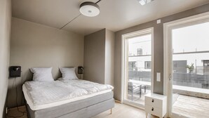 Une chambre moderne avec un lit, des lampes de chevet, un banc et une grande fenêtre donnant sur des immeubles.