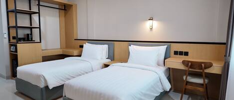 A hotel room with two beds, a desk, a chair, a shelf unit, and a wall-mounted light.