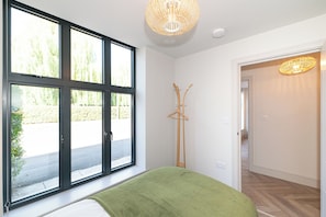 Ein Zimmer mit einem großen Fenster, einem Bett mit grüner Decke, einem Holzanziehkleiderständer und einem Flur, der in ein weiteres Zimmer führt.