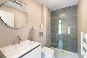 Ein modernes Badezimmer mit einem großen, ovalen Spiegel, einem weißen Waschbecken, einer Toilette und einer Duschkabine aus Glas.