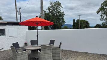 Eine Terrasse im Freien mit Tisch und Stühlen, einem roten Sonnenschirm und Blick auf Bäume und Berge.