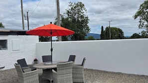 Een terras buiten met een tafel, stoelen, een rode parasol en uitzicht op bomen en bergen.